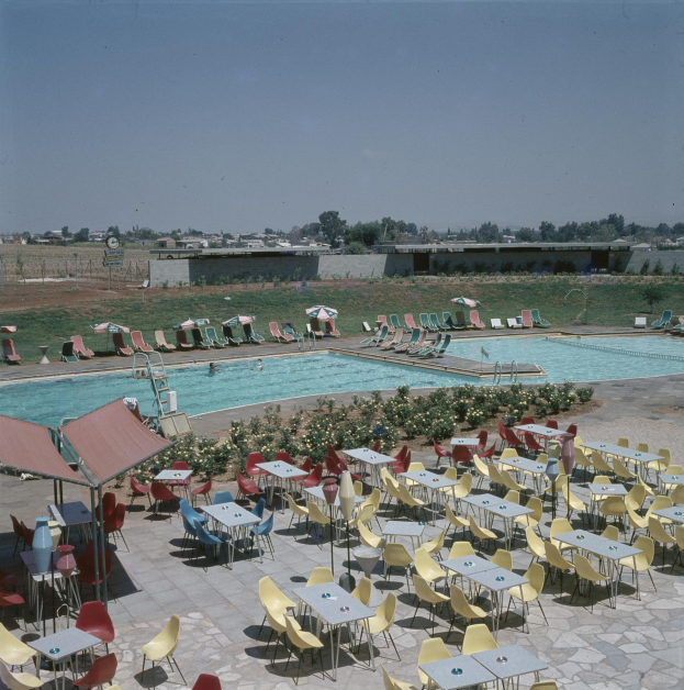 Ein Freibad mit Tischen, Stühlen und Sonnenschirmen, umgeben von Pflanzen, einem Zaun, einer Wand, Bäumen, Gebäuden und einer bewölkten Himmel.