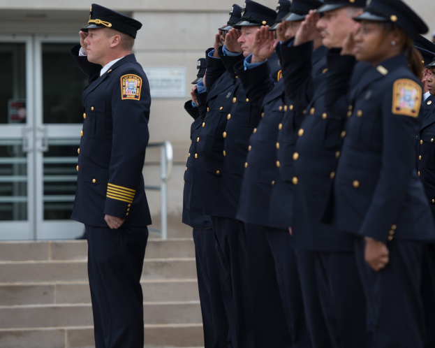 Eine Gruppe von Polizeibeamten in Uniform und Mützen steht in Formation vor einem Gebäude mit Glastüren und salutiert.