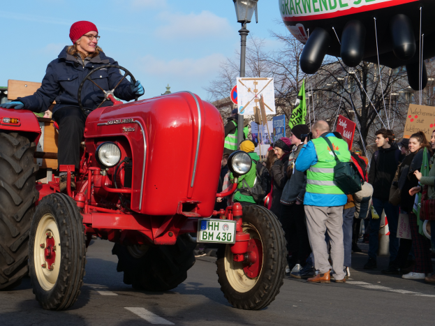 Eine Frau fährt einen roten Traktor auf einer Straße, umgeben von einer Menge mit Schildern und Fahnen, mit Laternen, Bäumen, Gebäuden und einem klaren blauen Himmel im Hintergrund.