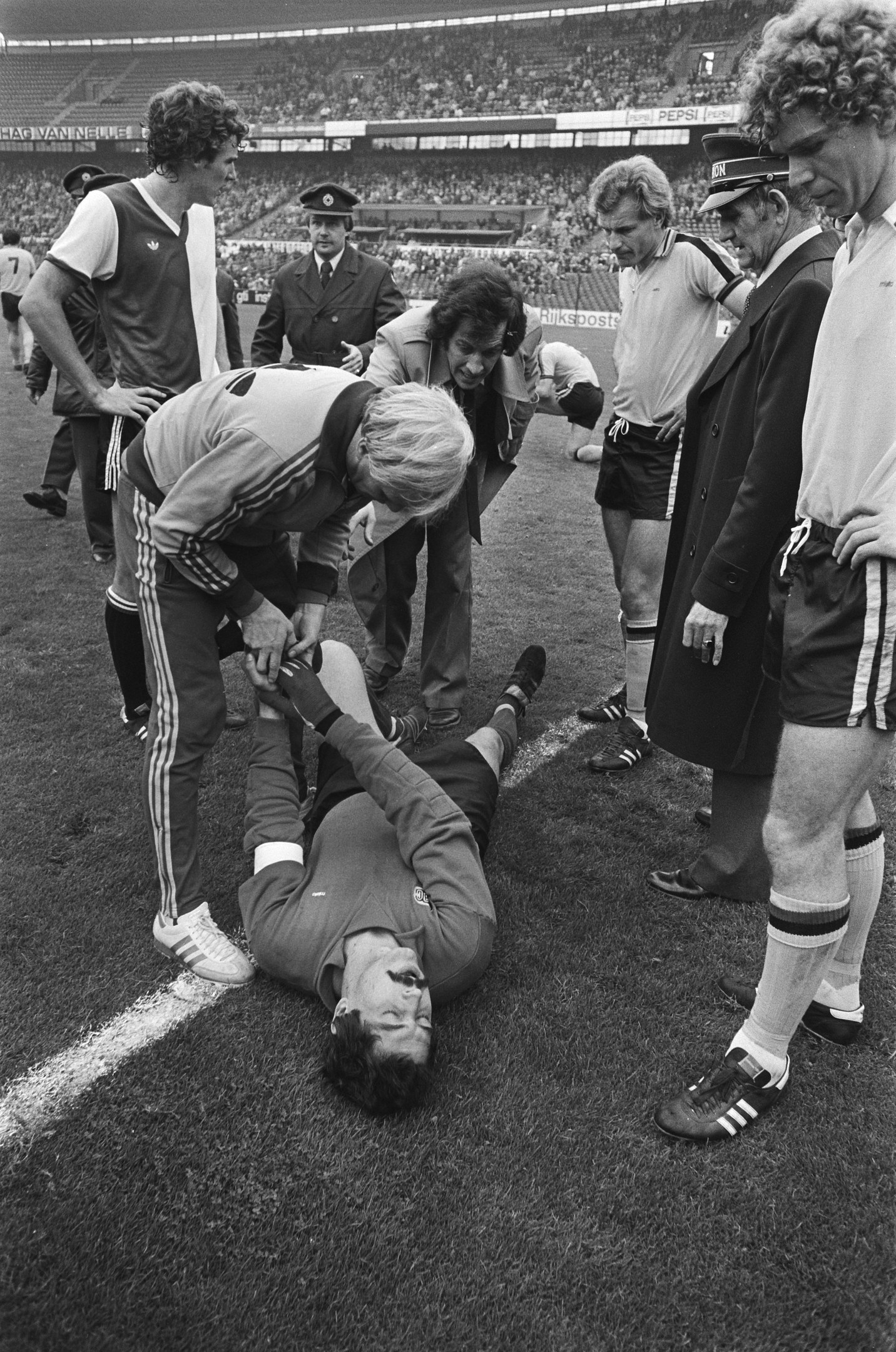 Schwarzes und weißes Bild eines Fußballspielers, der auf dem Boden liegt, während ihn eine Gruppe von Männern und ein Stadionpublikum umgibt; der Spieler scheint verletzt zu sein.
