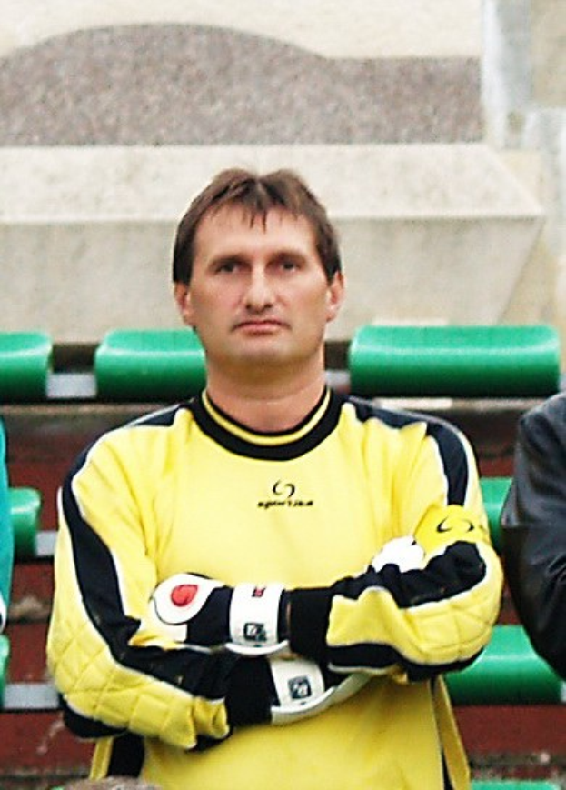 Ein Torwart in gelbem Trikot steht mit konzentrierter Miene neben einem anderen Mann in einer schwarzen Jacke, mit grünen Stühlen und einer Wand im Hintergrund.