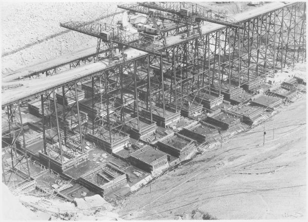 Ein Schwarz-Weiß-Foto einer Baustelle mit einer teilweise gebauten Metallbrücke, umgeben von ein paar Gebäuden, und Text unten.