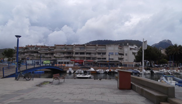 Ein Hafen mit Booten im Wasser, eine Brücke mit Geländern, Bänken, Laternenmasten, Bäumen, Gebäuden mit Fenstern, Hügeln im Hintergrund und einem bewölkten Himmel.