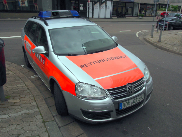 Ein Polizeiwagen steht am Straßenrand neben einem Mülleimer auf dem Gehweg, mit Gebäuden und Bäumen im Hintergrund.
