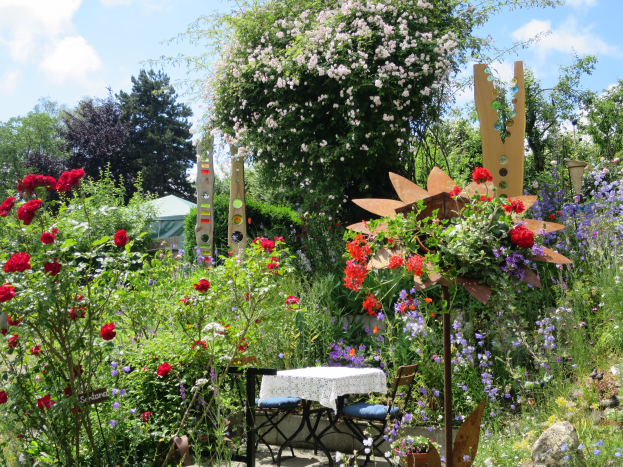 Ein Garten auf dem Chelsea Flower Show mit bunten Blumen und Pflanzen, einem Tisch mit einer Decke und Stühlen in der Mitte, umgeben von Bäumen unter einem bewölkten Himmel.