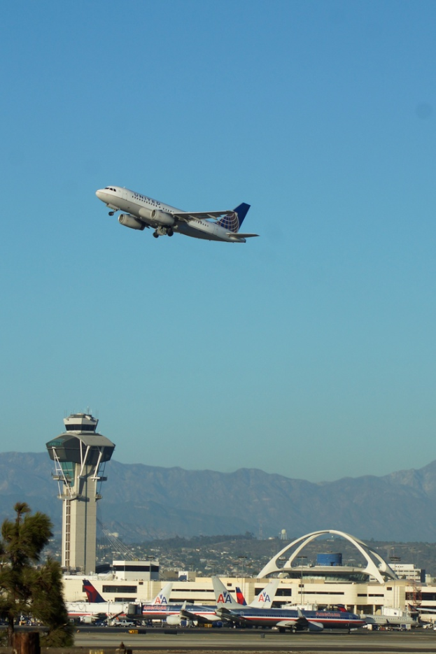 Ein Flughafenblick mit einem weißen Kontrollturm im Vordergrund, geparkten Flugzeugen am Boden, entfernten Gebäuden und einem Flugzeug am Himmel.