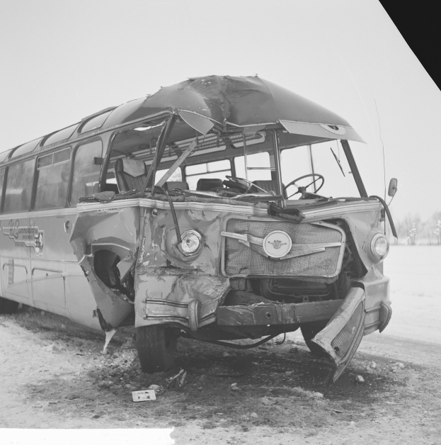 Ein Schwarz-Weiß-Foto eines schwer beschädigten Busses am Straßenrand mit einer zerquetschten Front, zersplitterten Fenstern, umgeben von Bäumen und einem bewölktem Himmel.