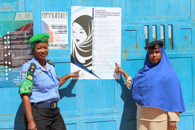 Zwei Frauen stehen nebeneinander vor einer blauen Tür mit Plakaten, eine in einem blauen Hemd, schwarzen Hosen und einer grünen Mütze, die andere hält ein Papier in der Hand.