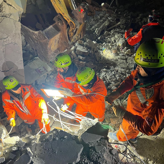 Eine Gruppe von Menschen in orangefarbenen Uniformen und grünen Helmen, die auf Schutt arbeiten, mit einem Kran im Hintergrund, der verschiedene Gegenstände in den Händen hält.