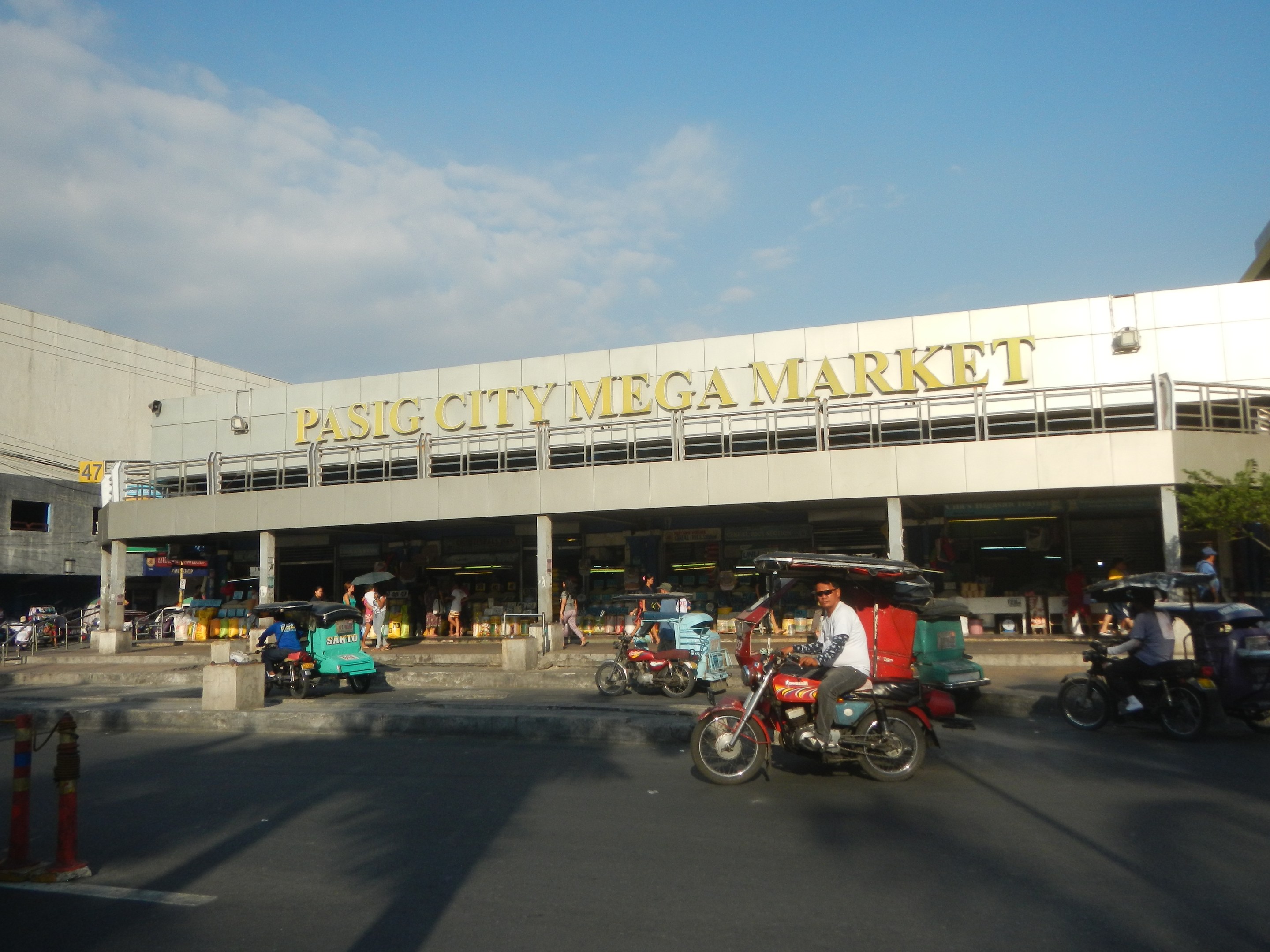 Eine belebte Straßenszene auf dem Pasig City Mega Market mit Fahrzeugen und Fußgängern, mit einem Gebäude mit Text im Hintergrund unter einem bewölkten Himmel.