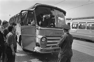 Schwarzes und weißes Bild einer Gruppe von Menschen, die um einen verunglützten Bus auf einer Straße mit anderen Fahrzeugen im Hintergrund stehen.