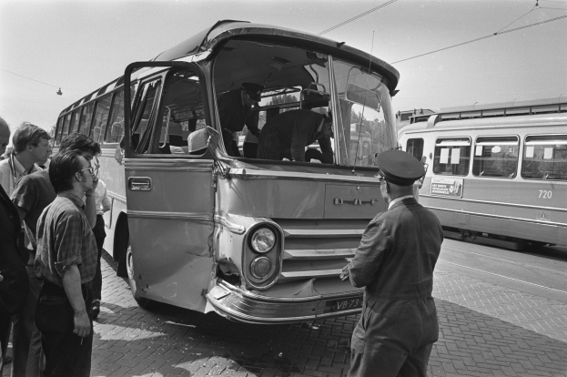 Schwarzes und weißes Bild einer Gruppe von Menschen, die um einen verunglützten Bus auf einer Straße mit anderen Fahrzeugen im Hintergrund stehen.