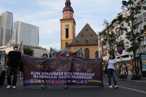 Eine Gruppe von Menschen mit Masken geht eine Straße entlang und hält ein Banner hoch, mit einem geparkten Auto auf der linken Seite, Gebäuden und Bäumen im Hintergrund und einem klaren blauen Himmel darüber.