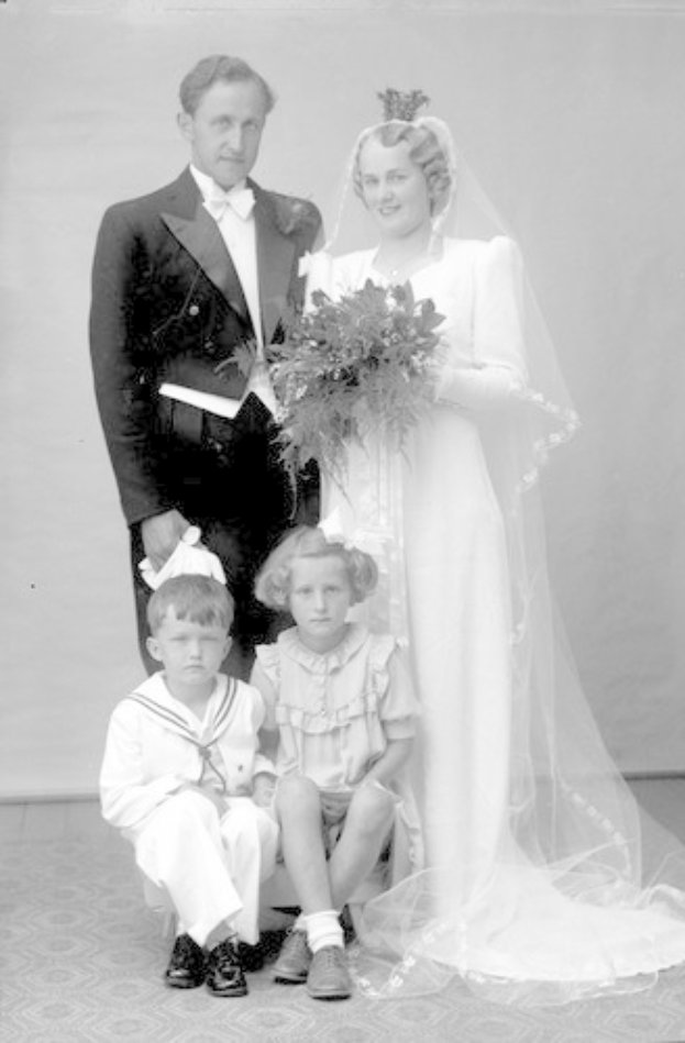 Schwarz-weiß-Foto einer Braut in einem weißen Kleid, die einen Strauß hält, neben ihr ein grinsender Bräutigam und zwei Kinder, die vor ihnen auf dem Boden sitzen.