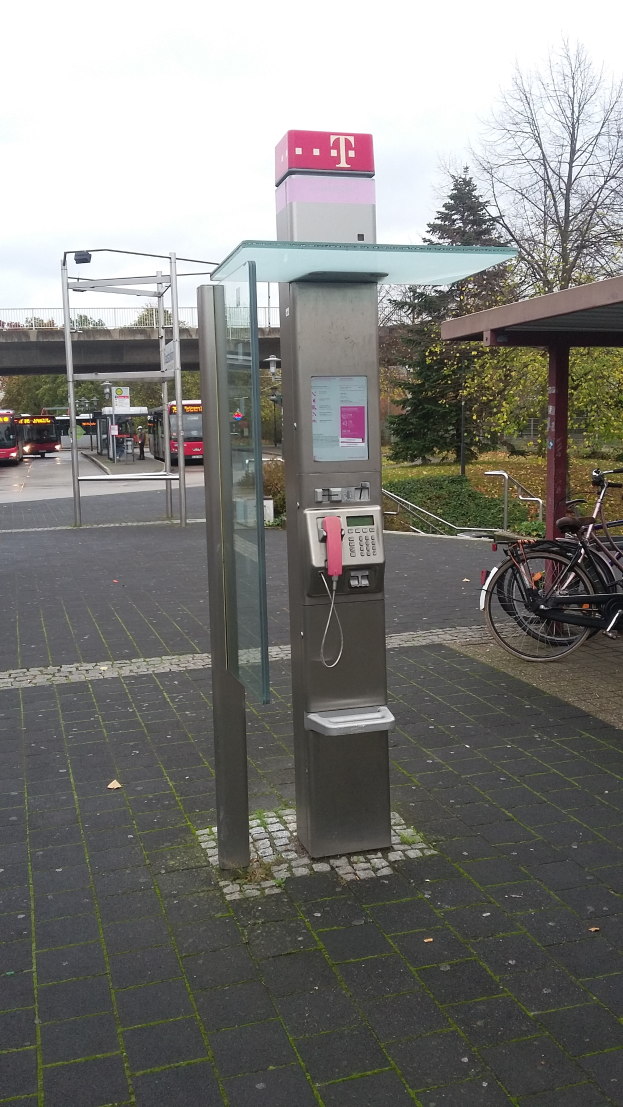 Ein Münzfernsprecher steht an einer Straße neben einer Bushaltestelle, daneben parkt ein Fahrrad; im Hintergrund sind Bäume, Fahrzeuge auf der Straße, eine Brücke und ein klarer blauer Himmel zu sehen.