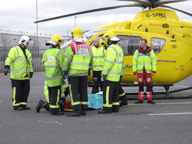 Eine Gruppe von Arbeitern in grünen Jacken steht auf einem Boden neben einem Hubschrauber, mit einem Zaun, kleinen Bäumen und einer Hütte im Hintergrund.