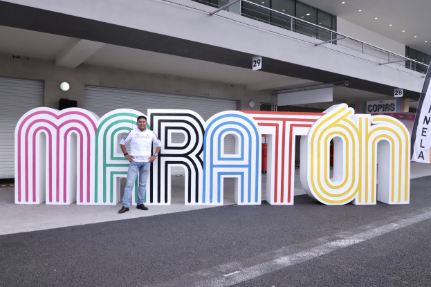 Ein Mann steht vor einem großen "Marathon"-Schild mit einem Banner rechts daneben, im Hintergrund ist ein Gebäude mit Lichtern, Geländern und Fensterläden zu sehen.