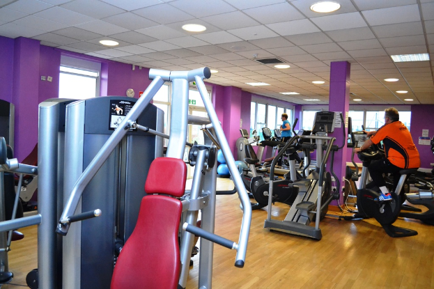 Ein Gym mit mehreren Geräten und einigen Menschen, die trainieren, mit einer Decke und Lampen oben im Bild.