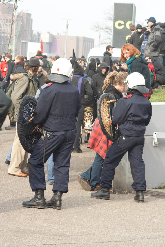 Eine Gruppe von Menschen, die auf einer Straße gehen, mit zwei Personen in der Mitte, die wie Polizisten aussehen, Gebäuden im Hintergrund und Boden unten.
