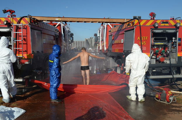 Drei Personen in hochvisiblen Westen stehen im Vordergrund, eine Person steht auf einer Abdeckung über Wasser, umgeben von ein paar anderen, Feuerwehrautos mit ausgefahrenen Metallstangenleitern und Gebäuden im Hintergrund.