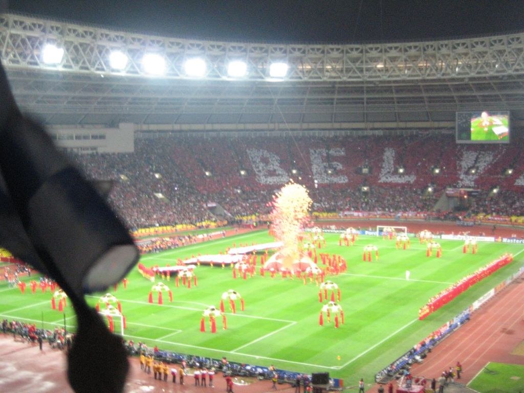 Ein Stadion mit Flutlicht, Eisengittern, Zuschauern, stehenden Personen, Werbetafeln, Anzeigetafeln, Überwachungskamera, Fahnen und Dekorationselementen.