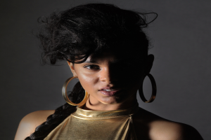 Eine Frau in einem goldenen Kleid und großen Ohrringen steht selbstbewusst vor einer Wand, ihr glatter Bob ist ordentlich frisiert, dezentes Make-up mit rosafarbenem Rouge und glänzenden Lippen, sie schaut direkt in die Kamera mit einem entschlossenen Gesichtsausdruck.