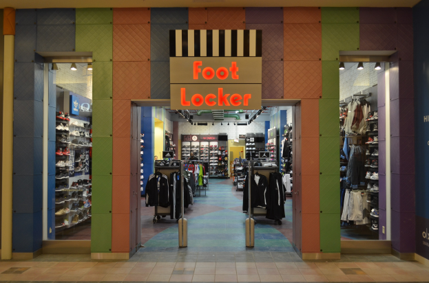 Innenansicht eines Foot Locker Schuhgeschäfts mit Kleidung an Bügeln und Regalen unter Deckenbeleuchtung.