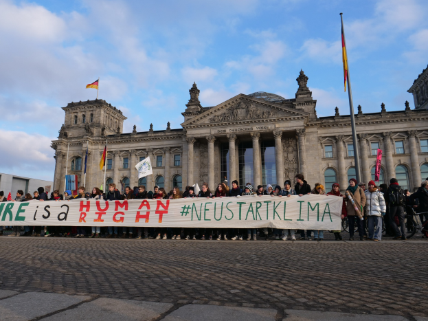 Eine Gruppe von Menschen hält ein Transparent mit der Aufschrift "Wir sind ein Menschenrecht" vor dem Reichstaggebäude in Berlin, Deutschland, mit seinen architektonischen Details und umgeben von Flaggen unter einem bewölkten Himmel.