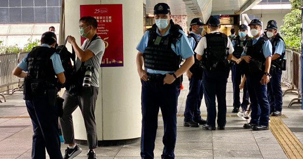 Eine Gruppe uniformierter Polizisten, einige mit Kameras, steht in einer Reihe vor einem Gebäude mit einem Banner an einer Säule, Geländern, Pflanzen, Bäumen und Deckenleuchten im Hintergrund.