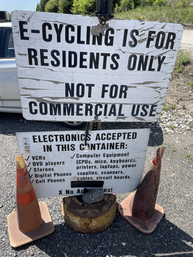 Sign reading "e-cycling is for residents only" placed on the roadside, bordered by traffic cones and other items, with a vehicle, trees, and a clear blue sky in the background.