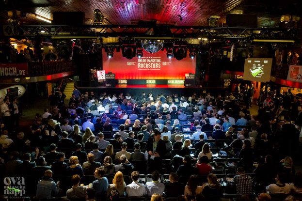 Eine große Menschenmenge sitzt auf Stühlen vor einer Bühne mit Bildschirmen, Bannern und Absperrungen, beleuchtet von Scheinwerfern, voller Erwartung auf ein Online-Marketing-Rockstars-Event, mit einem Wasserzeichen in der linken unteren Ecke.