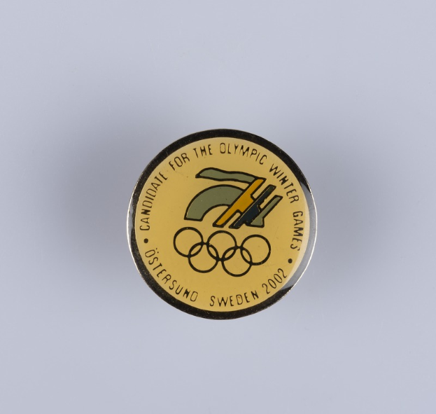 Ein runder Abzeichenaufnäher mit schwarzem Hintergrund und goldfarbenem Olympia-Logo in der Mitte, mit der Beschriftung "Olympische Winterspiele" in fetter weißer Schrift.