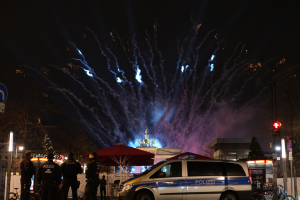 Ein Polizeiwagen steht vor einer Menschenmenge, umgeben von Fahrrädern, Zelten, Laternenmasten, Schildern, Verkehrszeichen, Bäumen, Gebäuden und einer Statue, unter einem von Feuerwerk erleuchteten Himmel.