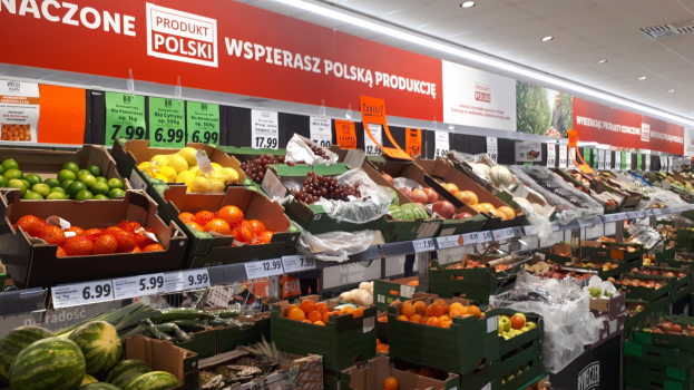 Ladeninneres mit Kartonschachteln verschiedener Obst- und Gemüsesorten, jeweils mit Preisschildern, Text- und Bildtafeln an den Wänden und Deckenleuchten.