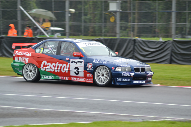 Ein Auto auf einer Rennstrecke mit Text und Zahlen, umgeben von Gras, mit Maschendrahtzaun, schwarzem Tuch, Menschen in orangefarbenen Jacken mit Schirmen und Bäumen im Hintergrund.