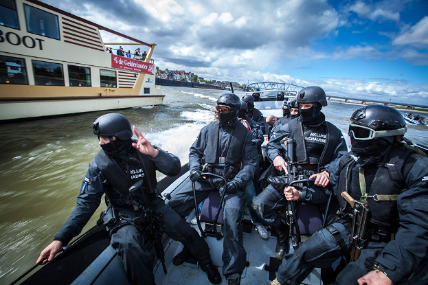 Eine Gruppe von Polizisten in schwarzen Uniformen und Helmen, einige bewaffnet, auf einem Boot im Wasser mit einer Brücke, Bäumen, Gebäuden und einem klaren blauen Himmel im Hintergrund.