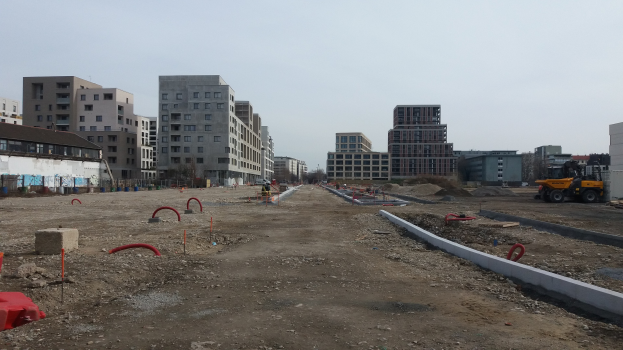 Baustelle mit schwerer Maschinerie im Vordergrund, Gebäude und Bäume im Hintergrund unter klarem Himmel.
