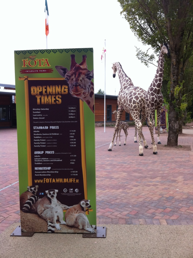 Giraffen vor Bäumen, Hütten und Flaggen mit einer Tafel, die Text, Logos und Tierbilder zeigt.