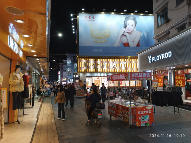Eine nächtliche Stadtstraße mit Menschen, die gehen, Gebäuden auf beiden Seiten mit Schaufensterpuppen in Kleidern, einem Stand mit einer Person auf einem Roller und verschiedenen Gegenständen und einem Plakat mit einem Bild einer Frau.