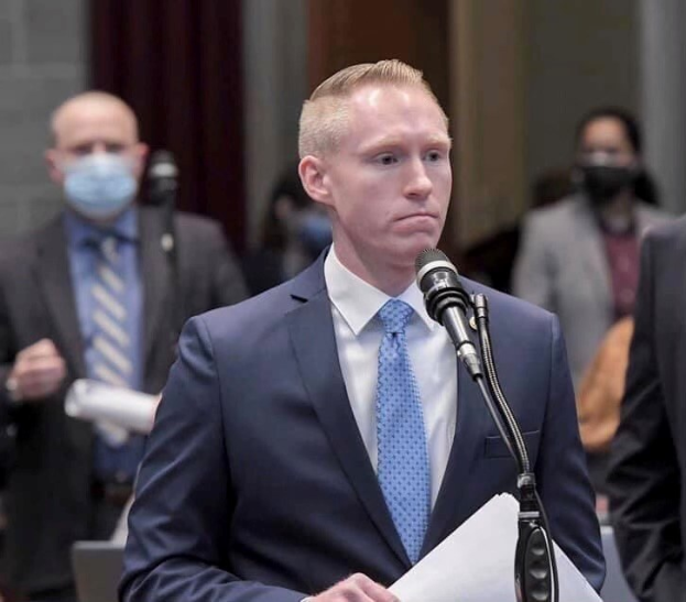 Ein Mann in Anzug und Krawatte spricht an einem Podium mit Papieren, gegenüber einem Mikrofon, während andere in der Nähe stehen und eine Vorhangwand im Hintergrund zu sehen ist.