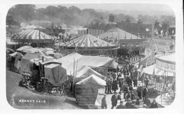 Ein altes Schwarz-Weiß-Foto einer belebten Karnevals-Szene mit Zelten, Menschen, Karren, Pfählen, Bäumen und Gebäuden im Hintergrund, mit dem Text "Barnet Fair, London, England" unten.