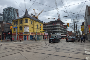 Eine Straßenszene in der Stadt mit einem gelben Gebäude im Bau in Vordergrund, Autos und Fußgänger auf dem Gehweg, Strommasten mit Drähten, Ampeln, Gebäude mit Fenstern, Schilder, Bäume, Kräne und ein bewölkter Himmel.