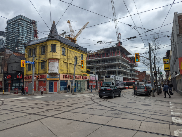 Eine Straßenszene in der Stadt mit einem gelben Gebäude im Bau in Vordergrund, Autos und Fußgänger auf dem Gehweg, Strommasten mit Drähten, Ampeln, Gebäude mit Fenstern, Schilder, Bäume, Kräne und ein bewölkter Himmel.