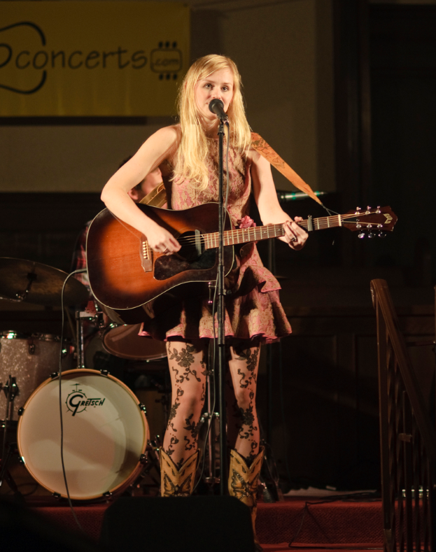 Eine Frau spielt Gitarre und singt auf der BÃ¼hne bei einem Konzert, mit einer Band und einem Bildschirm im Hintergrund und einer TribÃ¼ne unten.