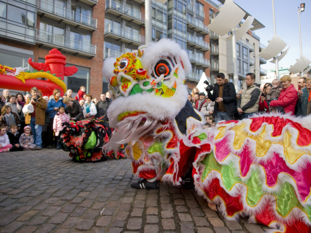 Ein lebendiges chinesisches Neujahrsfest in Amsterdam mit einer Löwen-Tanz-Vorführung vor einer Zuschauermenge, einige halten Kameras, vor einem Hintergrund aus Gebäuden, Laternenmasten und einem klaren blauen Himmel.