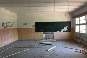 Ein leerer Klassenraum mit graffiti-beschmierten Wänden, einer zentralen Tafel, gebrochenem Holz auf dem Boden und großen Fenstern auf der rechten Seite.