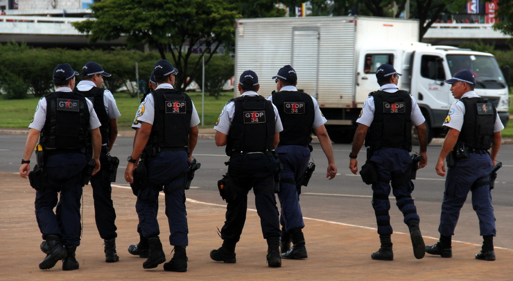 Eine Gruppe von uniformierten Beamten, einschließlich Mützen, Hemden, kugelsicheren Westen, Uhren, Hosen und Schuhen, die auf einer Straße mit einem geparkten Lastwagen auf der rechten Seite neben einem Zaun, Gebäuden, Bäumen, Pflanzen, Pfählen und Bannern im Hintergrund und einer Brücke im oberen rechten Eck gehen.