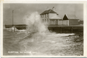 Eine große Welle kracht gegen eine Mole in einem Schwarz-Weiß-Foto, mit Häusern, Pfählen und einer Brücke im Hintergrund und Text unten.