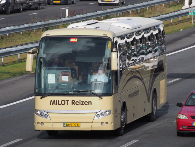 Ein Minibus mit Passagieren fährt auf einer Autobahn neben einem roten Auto, mit Leitplanken, Gras und textbehafteten Säulen im Hintergrund.