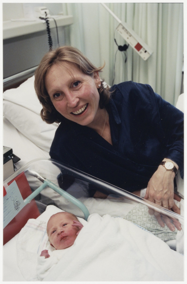 Eine Frau in einem blauen Kleid und einer Uhr wiegt ein Baby in ihren Armen, während sie in einem Krankenhausbett liegt, umgeben von medizinischer Ausrüstung und einem weißen Vorhang.